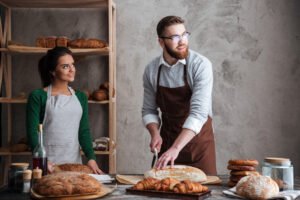 Picture of cheerful loving couple bakers. Man cut the bread. Looking aside.